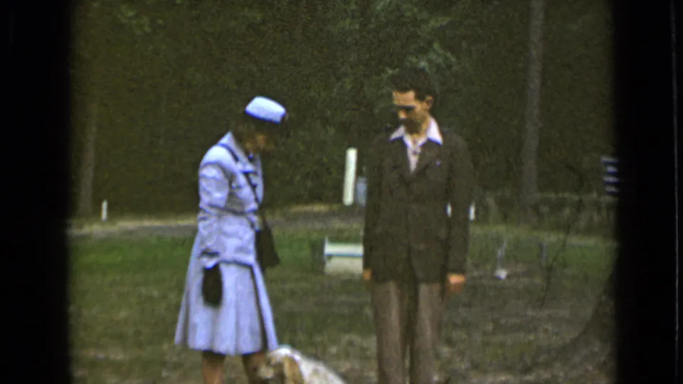 The couple stands near the small dog in the supported 1947 New York park footage, framed by grass, trees, and soft 8mm color.