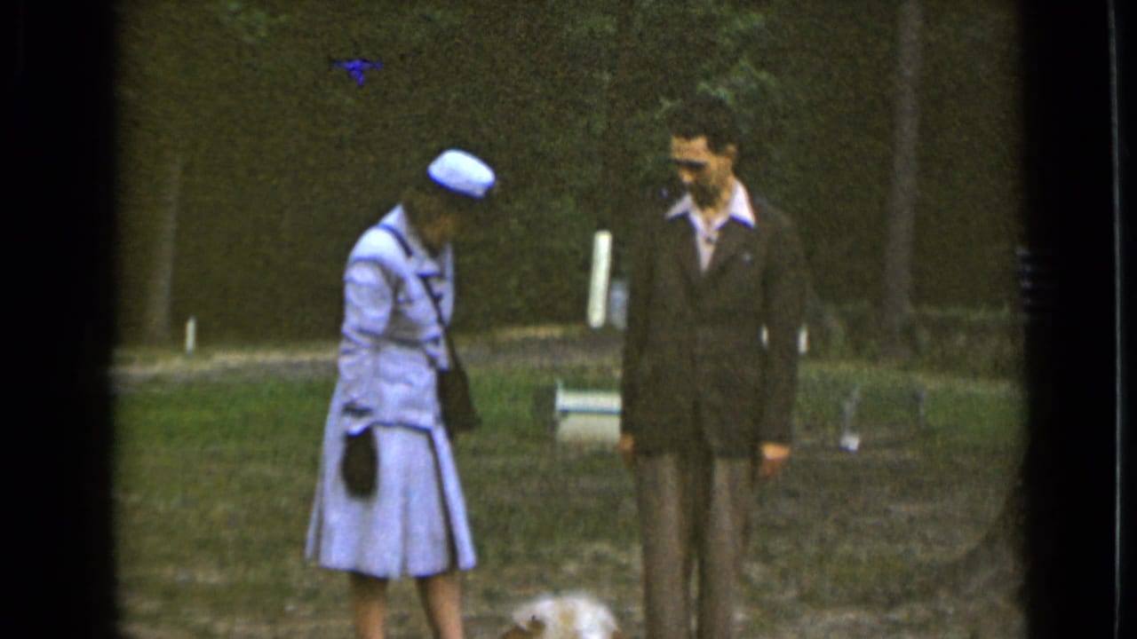 In 1947 New York home-movie footage, a woman in a pale coat and hat stands beside a suited man while a small dog is at their feet.