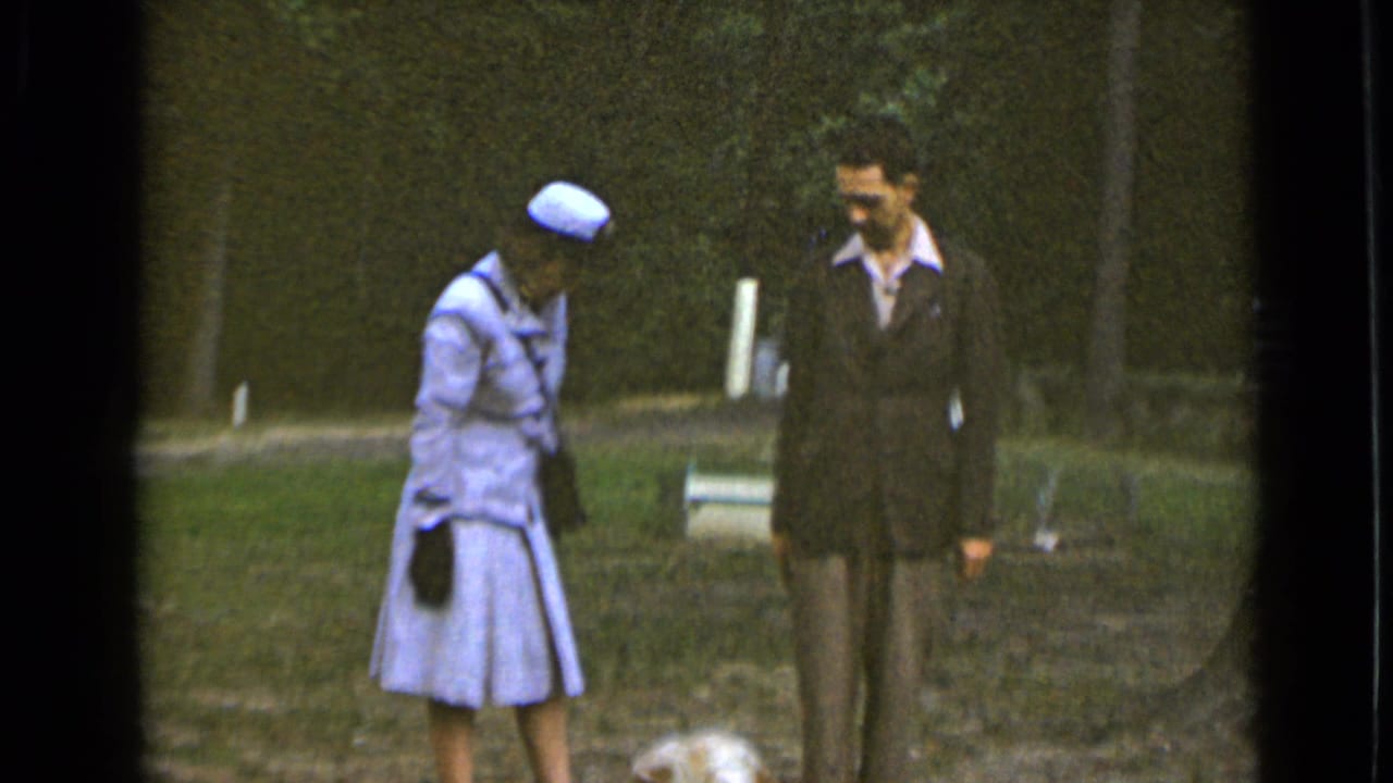 Woman and suited man stand near a small dog on grass with trees behind them.