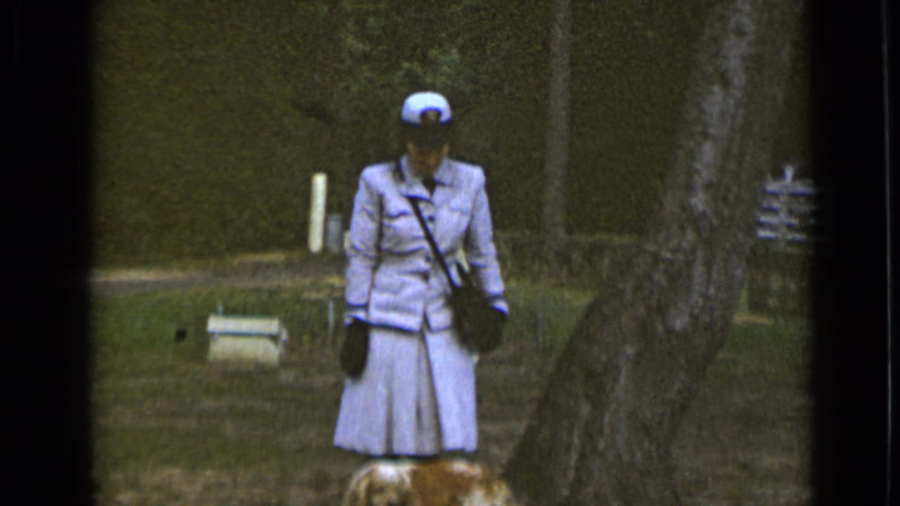 In 1947 New York home-movie footage, a woman in a light uniform-style outfit stands by trees with a small dog at her feet.