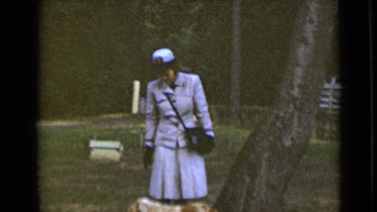 Woman in a cap looks down while standing near a small dog and trees.