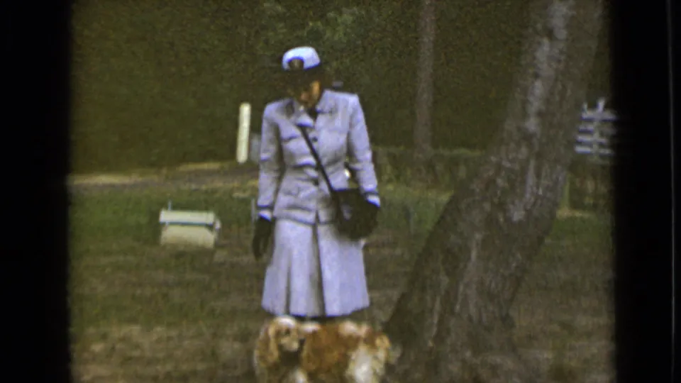 A woman in a light uniform-style outfit stands near a small dog at the end of the short 1947 New York park clip.
