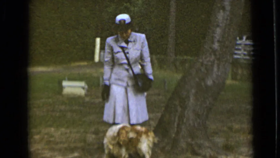In 1947 New York home-movie footage, a woman in a light official-style uniform stands with a golden spaniel in a grassy park area.