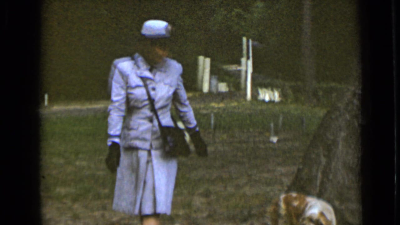 Woman in a pale blue suit and hat walks on grass with a small dog near her feet.