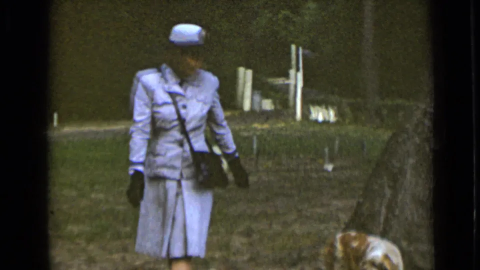 In 1947 New York home-movie footage, a woman in a pale blue uniform-style suit and hat walks through a grassy park with a small dog nearby.