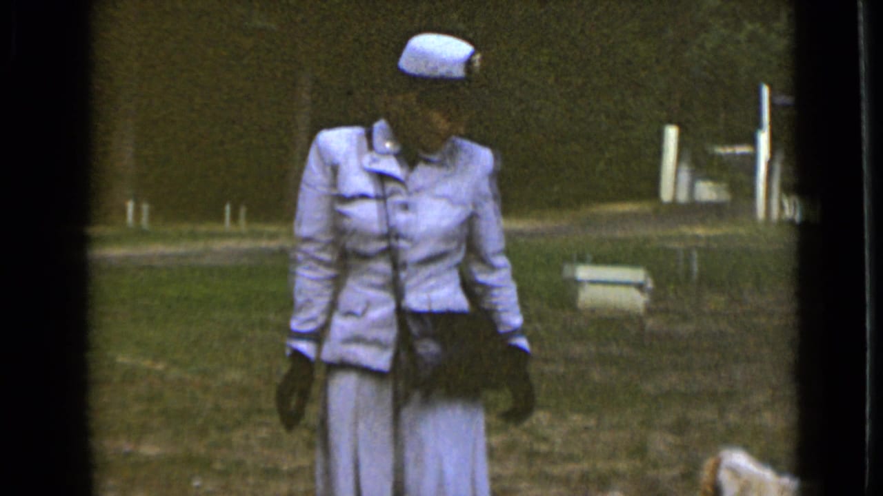 Woman in pale blue clothing stands or walks on grass with a small dog partly visible at lower right.