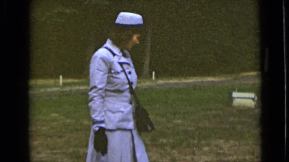 Side view of the woman in a pale blue suit, matching hat, gloves and shoulder purse walking across grass in 1947 New York footage.