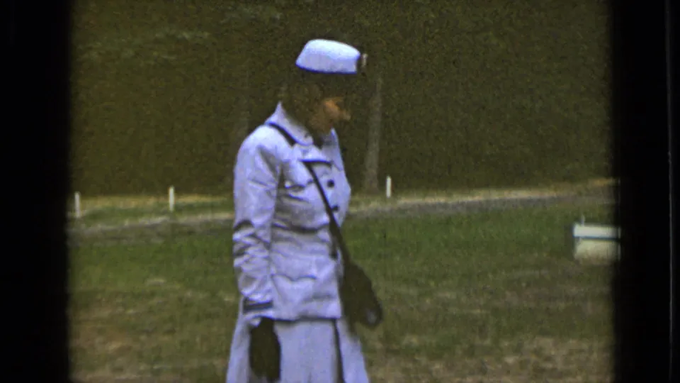 The woman remains in profile, wearing a pale blue suit, matching hat, dark gloves and purse while walking through a grassy park area.