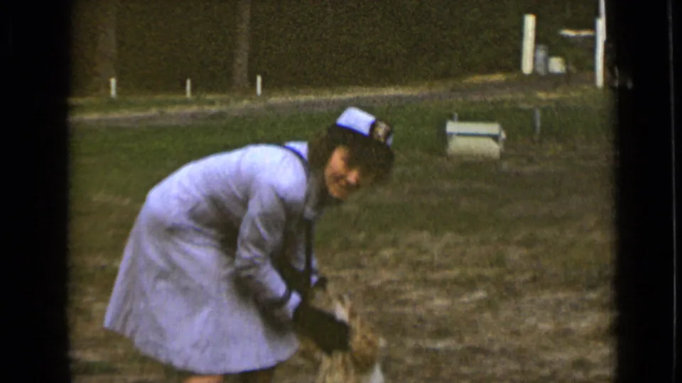 The woman bends and smiles as she reaches toward a long-haired dog in the grassy field, with trees and a small bench visible behind her.
