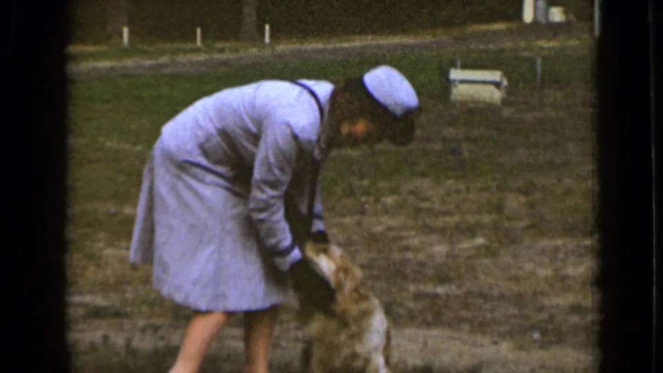 The woman leans over the long-haired Cocker Spaniel in the field, appearing to pet or steady the dog during the brief home-movie moment.