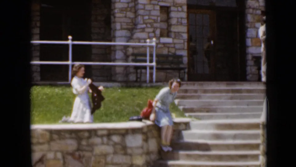 Two women in 1940s attire outside a stone-fronted cavern site, with steps, railings, grass, and a building entrance visible.