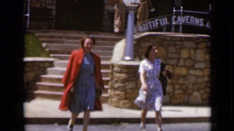 Two women in dresses walk past a stone wall and steps, with a partially visible Beautiful Caverns sign overhead.