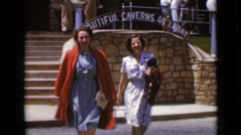 Two women approach the camera outside the cavern attraction, with stone steps, a lamp post, and the curved sign behind them.