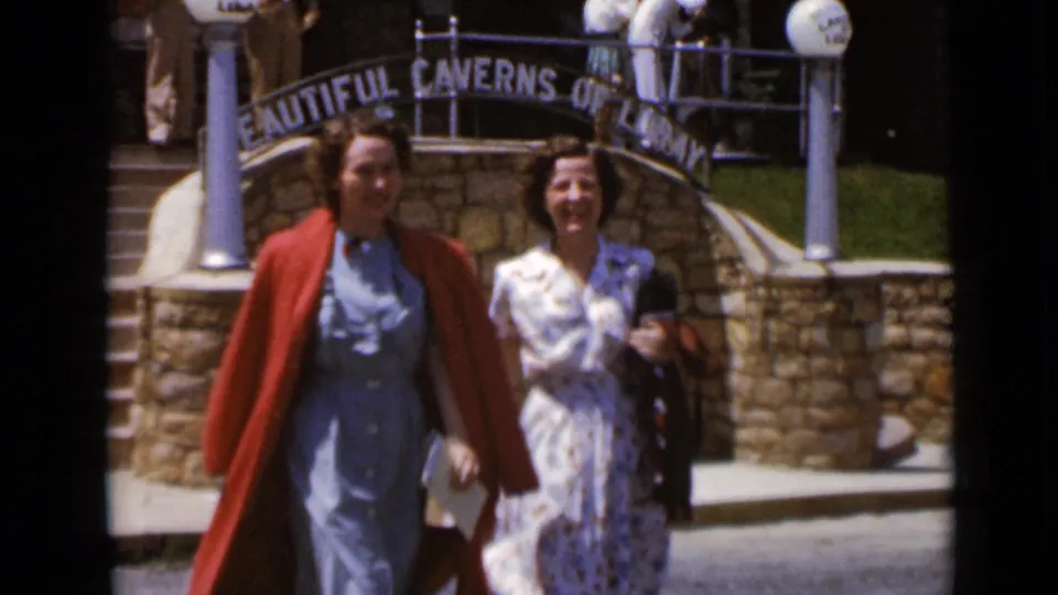 Two women in 1940s attire walk together near a stone arch bearing Caverns of Luray signage.