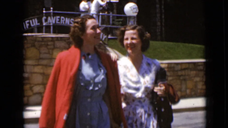 Two smiling women in vintage dresses walk arm-in-arm past a stone entrance with Caverns signage behind them.