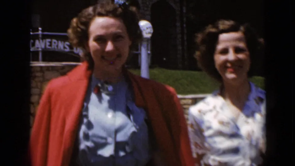 Close 8mm frame of two women smiling near a Caverns entrance sign, with one wearing a red coat over a blue dress.