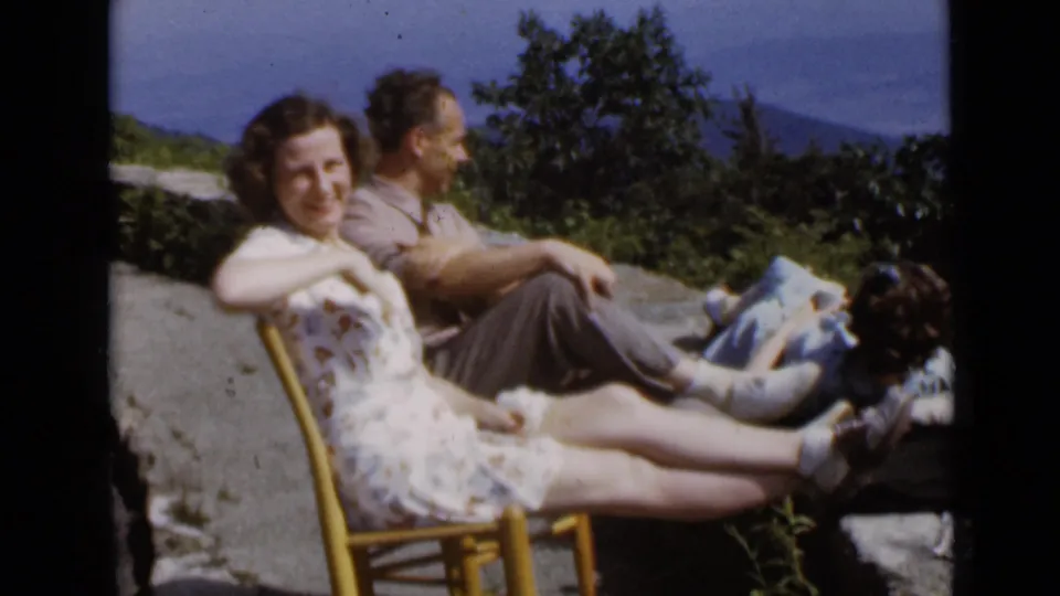 A woman and man relax on a hillside or overlook, with another reclining figure partly visible and mountains in the background.