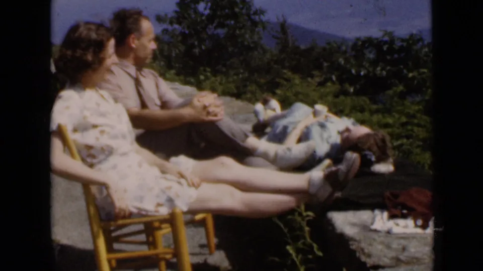 1947 New York home-movie frame showing two women seated outside and a man lying in the sun near trees and distant hills.