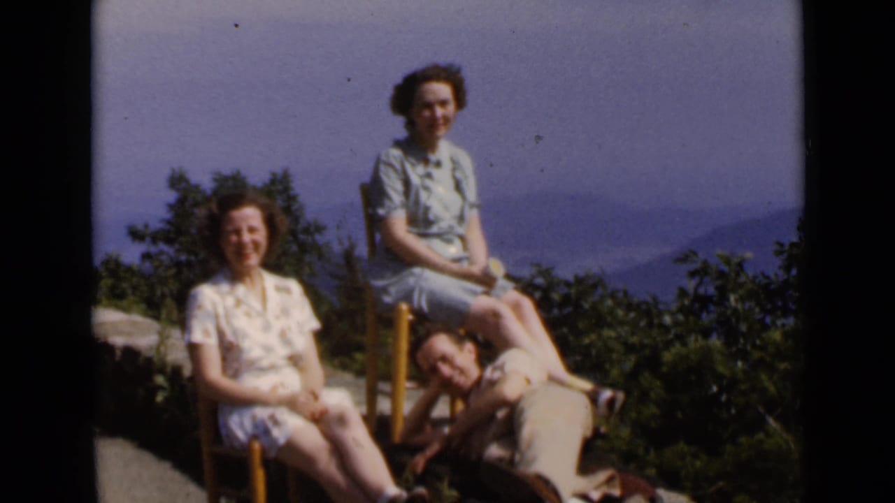 Two women sit in outdoor chairs as a man rests with his head near their feet.