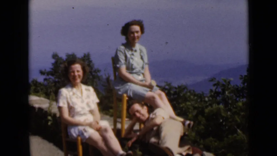 1947 New York frame with two women seated on chairs and a man reclining at their feet against trees and hazy hills.