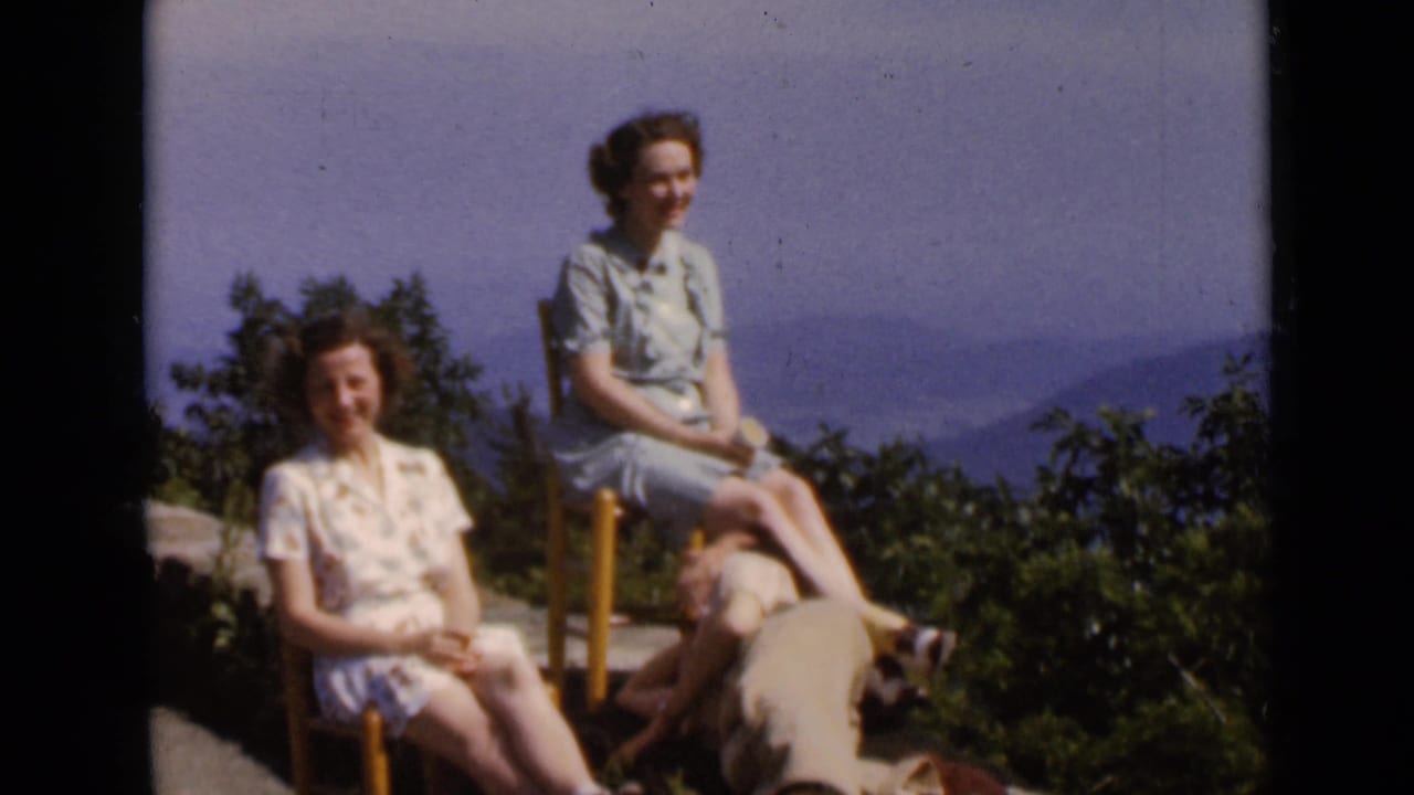 1947 New York home-movie frame showing two women seated at a mountain overlook with a man reclining nearby and hazy hills behind them.