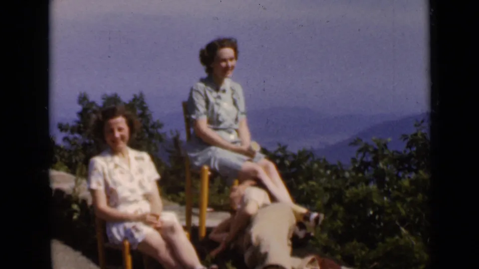 1947 New York home-movie frame showing two women seated at a mountain overlook with a man reclining nearby and hazy hills behind them.