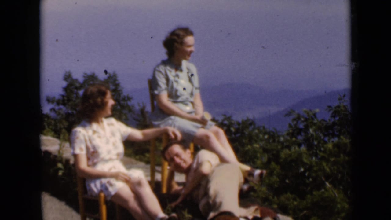 Two women sit outdoors at an overlook as a man leans on the ground beside them.