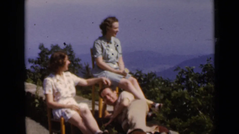 1947 New York home-movie frame with two women in light dresses seated outdoors and a man leaning near them at a wooded mountain overlook.