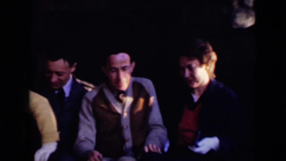 Dim 1947 home-movie interior showing three men seated close together, including a man in a vest and tie at center.