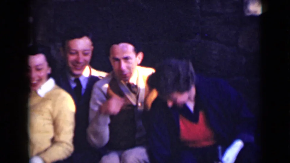 1947 New York home-movie frame showing adults in period clothing seated closely in a dark stone-walled interior.