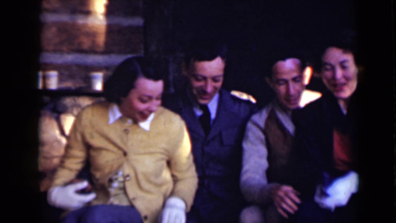 Four adults sit close together near a stone fireplace, smiling and looking down.