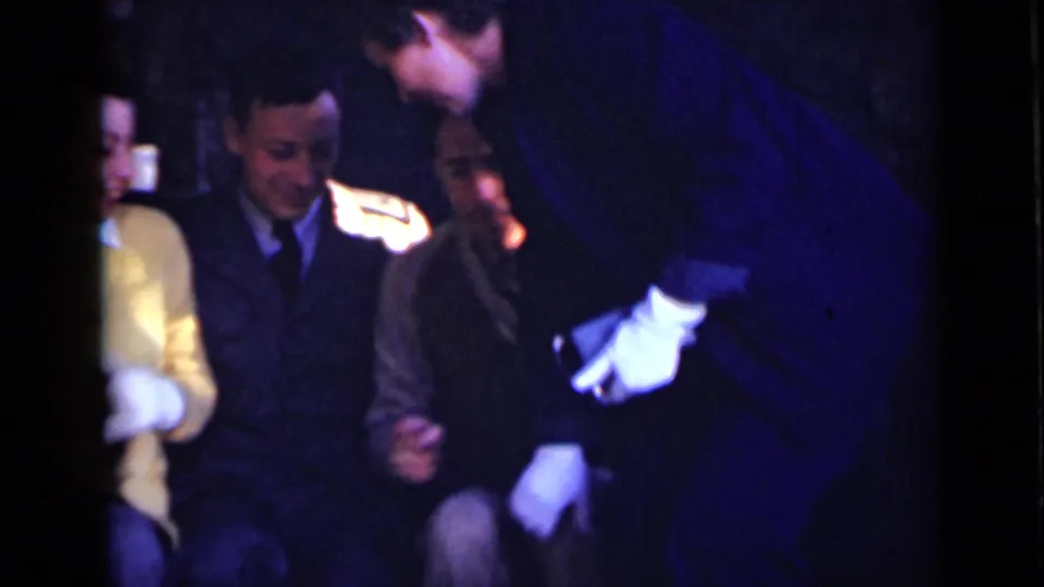 A 1947 New York home-movie frame shows a seated group partly obscured as one adult leans into the foreground.