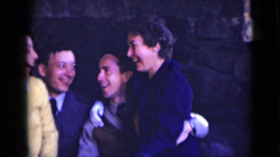 In this 1947 New York home-movie frame, adults lean close and laugh in a rustic stone-walled interior.