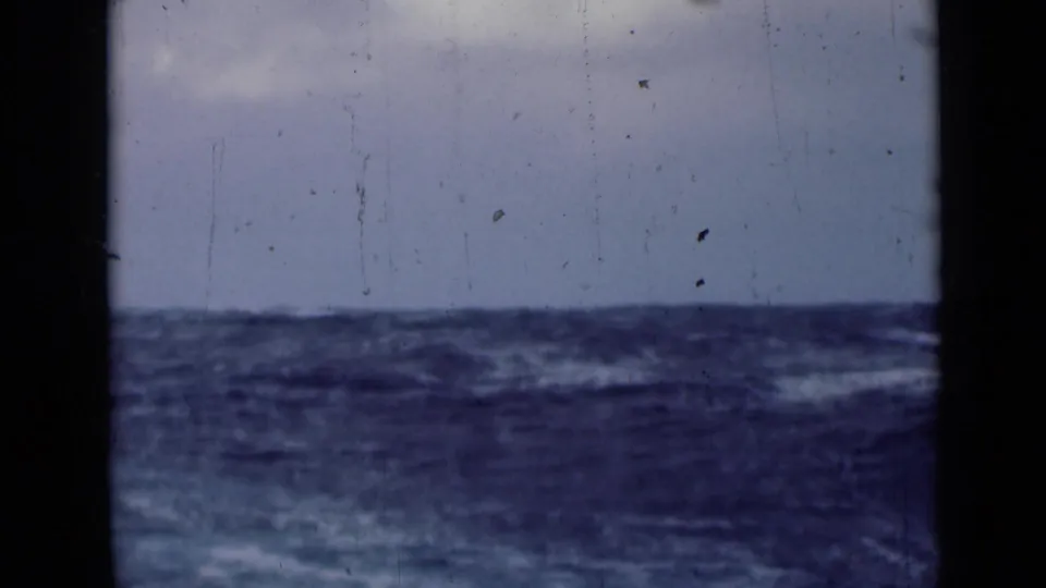 Rough open water and a low horizon under overcast sky, filmed from a moving vessel in archival 8mm footage.