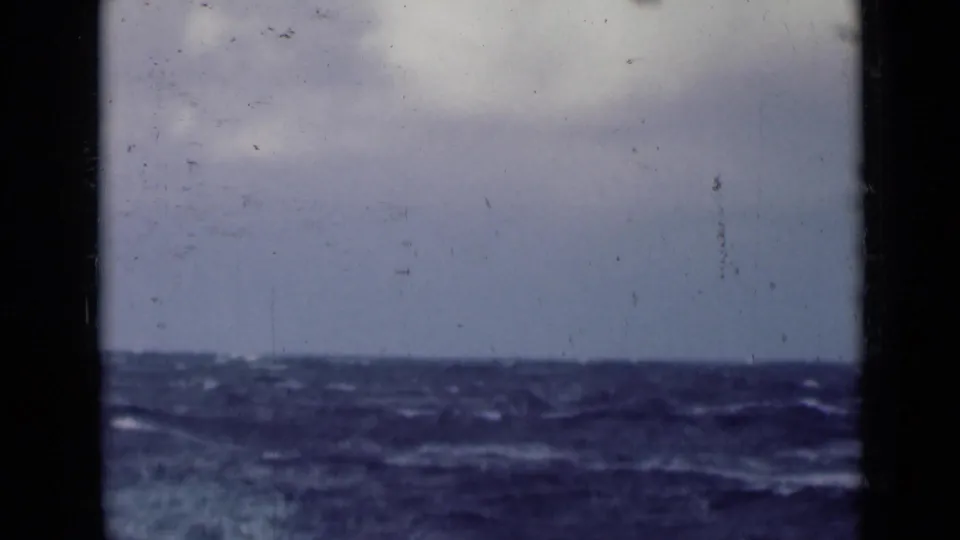 A low horizon of rough sea beneath a pale cloudy sky, with 8mm grain and dust visible across the frame.