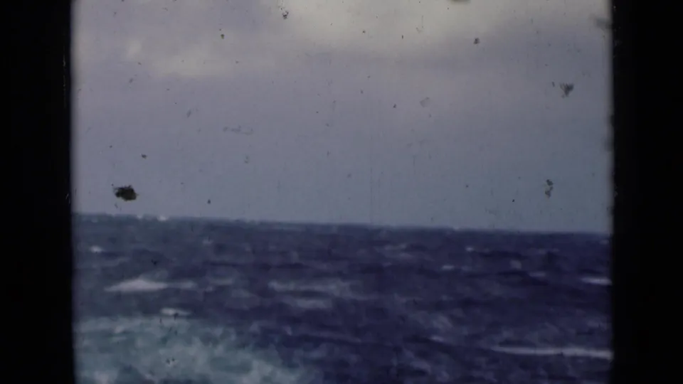 Open sea with rolling waves and whitecaps fills the lower frame, with overcast sky above in a faded 8mm scan.