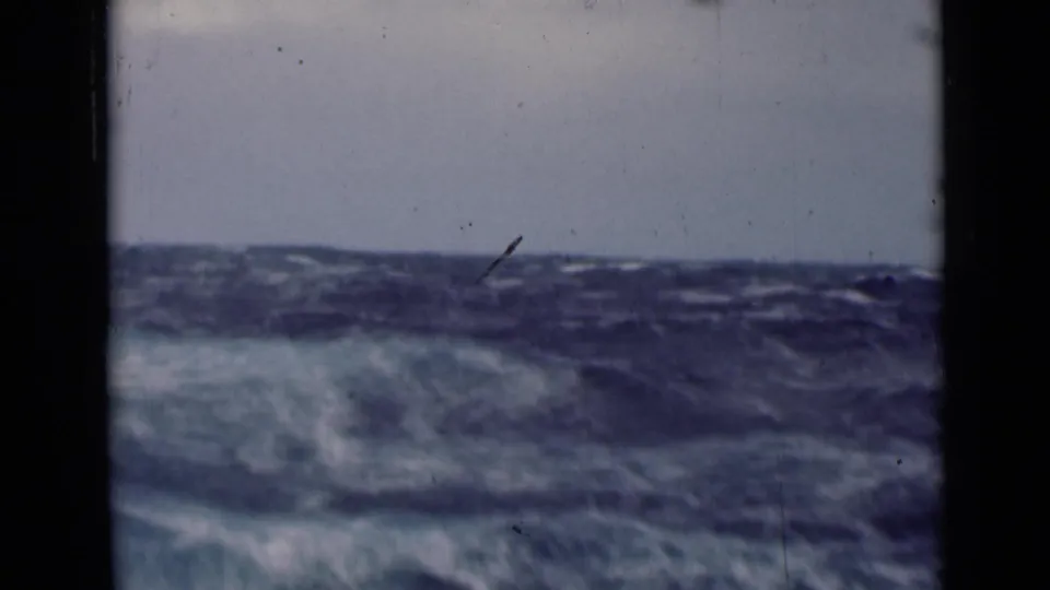 Rough open water rolls across the frame in a vintage 8mm shipboard shot, with scratches and heavy gate edges visible.