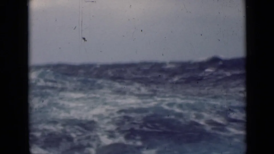 Vintage 8mm view of agitated blue sea and foam from a moving vessel, with soft focus and visible film wear.