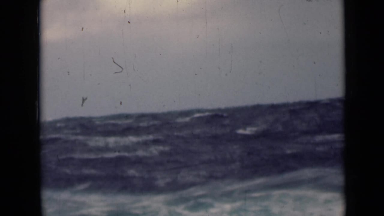 Rough open-water swells fill the lower frame beneath an overcast sky, with soft 8mm texture, dust, and dark gate edges visible.