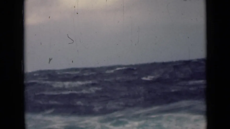 Rough open-water swells fill the lower frame beneath an overcast sky, with soft 8mm texture, dust, and dark gate edges visible.