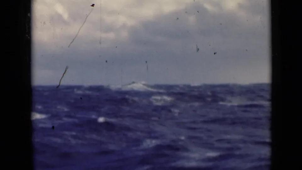 A choppy sea surface rises toward a low horizon beneath muted clouds, with scratches and dust marking the 8mm home-movie frame.