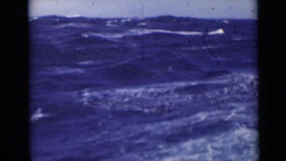 A larger swell crosses the frame with foamy streaks on dark blue water and visible scratches in the film image.