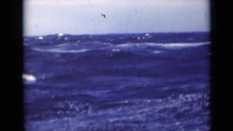 Rolling open sea with scattered whitecaps below a pale sky, shown through faded 8mm color and dark frame edges.