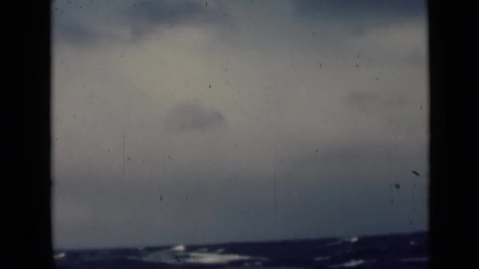 Faded 8mm frame of dark sea under a hazy, clouded sky, with the horizon low and no visible people or structures.