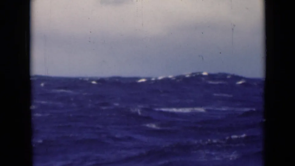 A passenger-view frame of rough open sea, showing blue wave crests, a pale sky, and the dark edges of scanned 8mm film.