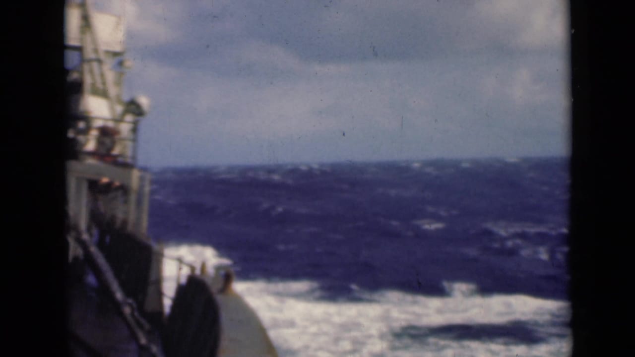 Boat side and deck edge beside rough blue water and white foam under a bright cloudy sky.