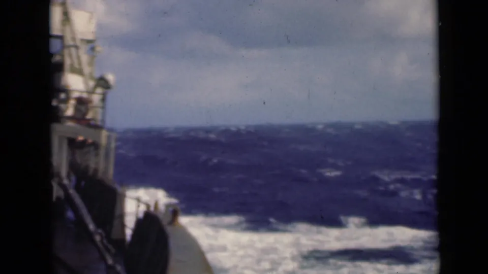 1940s 8mm frame from a moving vessel, with railings, deck edge, and churning open water visible under an overcast sky.