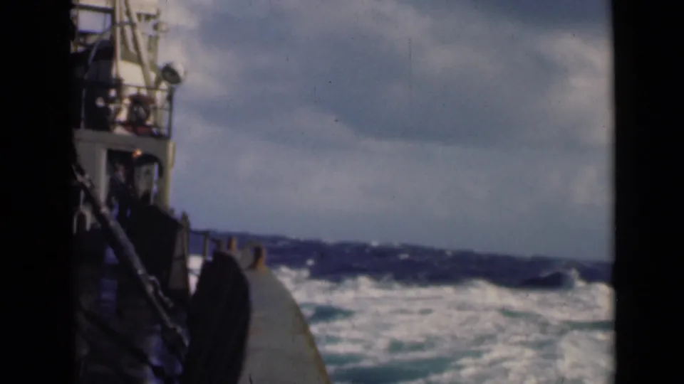 Vintage 8mm shipboard frame showing a partial vessel side, deck edge, railings, and foaming rough sea beneath a bright overcast sky.
