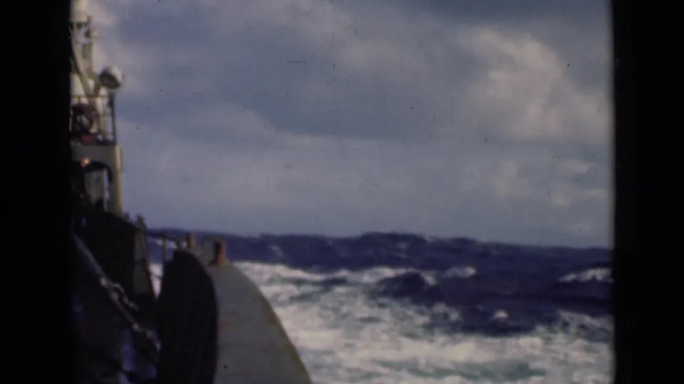 8mm home-movie frame from aboard a vessel, showing the boat edge against large dark waves and white spray under clouded daylight.