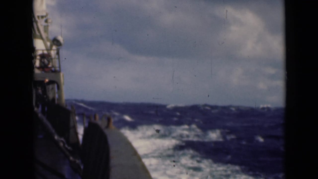 Deck-level view from a vessel cutting through rough seas, with railings, wake, whitecaps, and cloudy sky visible.